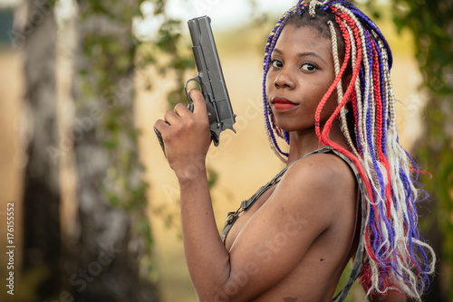 portrait of a beautiful young African American girl with a dreddy hairdress on a park background, nature, deciduous forest, with a gun in her hands. concept of photo walks, female self-defense