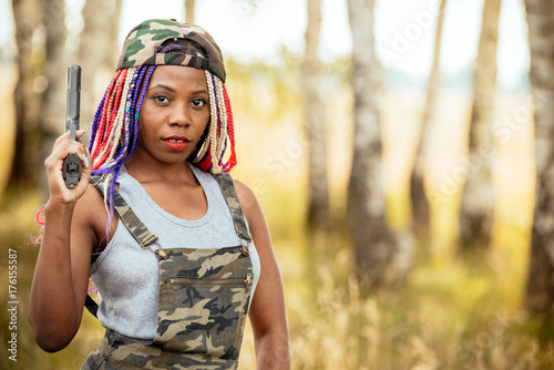 portrait of a beautiful young African American girl with a dreddy hairdress on a park background, nature, deciduous forest, with a gun in her hands. concept of photo walks, female self-defense