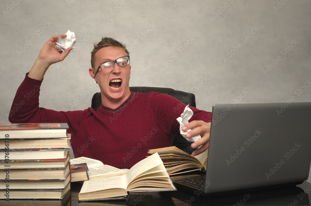Foto Stock Young student man becomes hysterical from studying. Office  worker goes crazy with overload at work. Mad student sitting at the table  and throws the piece of paper and prevents to
