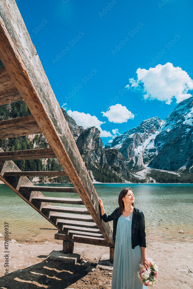 Beautiful young brunette woman posing near wooden pier near the lake in blue dress with wreath in hand. at lake, lago di braies,Dolomite,Italy.  have fun outdoor in summer stylish lifestyle portrait 
