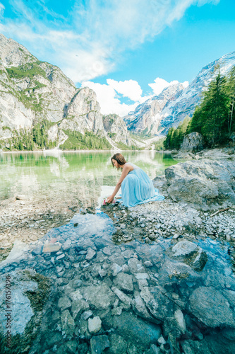 Woman in long blue dress stay on stone near braies lake in south tyrol, italy. Reflection in the water.