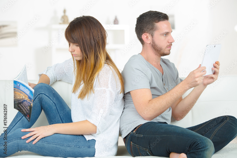 Couple sitting back to back reading book and tablet screen Couple sitting back to back reading book and tablet screen