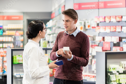 Experienced female pharmacist checking the indications and contraindications of a new medicine next to a young male customer in a modern pharmacy