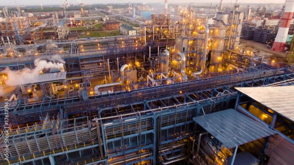 A huge oil refinery with metal structures, pipes and distillation of the complex with burning lights at dusk. Aerial view