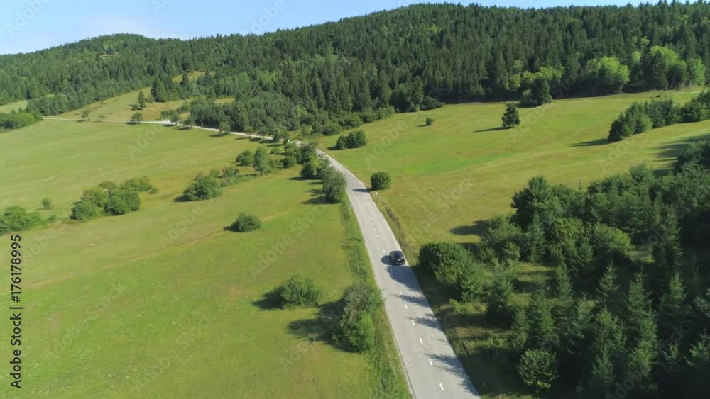 AERIAL: Black SUV car driving along empty country highway in idyllic rural landscape. People on road trip journey across beautiful green Slovenia. Lush green hills and meadow fields in remote valley AERIAL: Black SUV car driving along empty country highway in idyllic rural landscape. People on road trip journey across beautiful green Slovenia. Lush green hills and meadow fields in remote valley