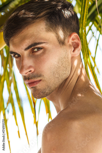 Handsome mature male model standing topless under palms on the beach, thinking while his eager eyes searches for the ray of sun, shadows falling on his body.
