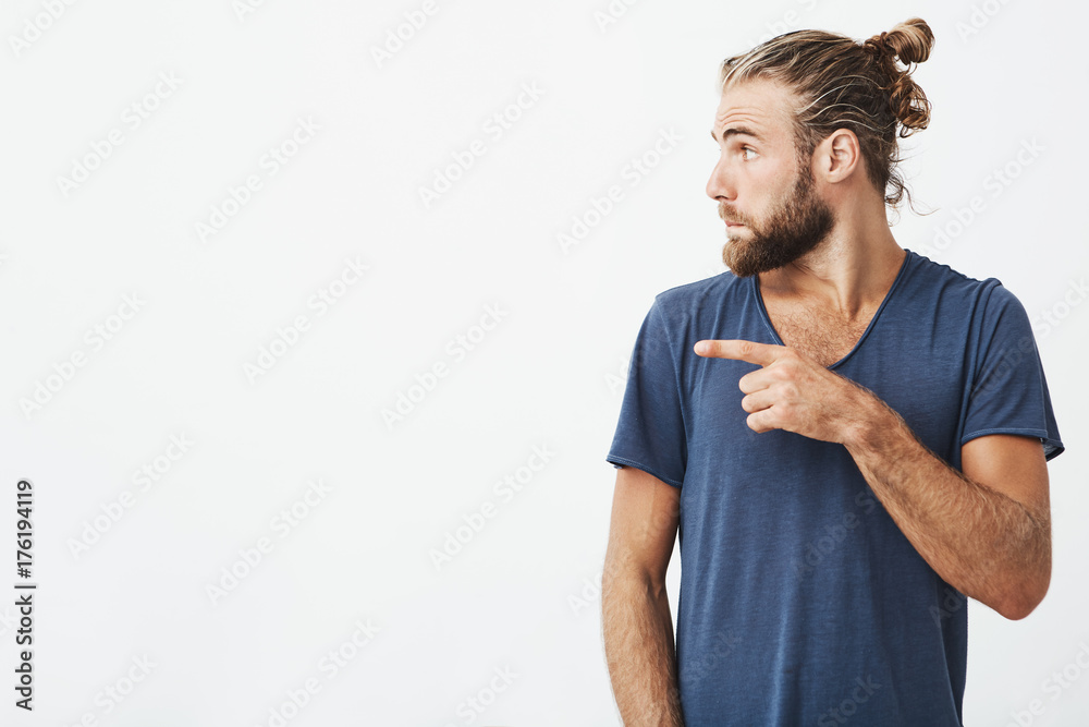 Close up portrait of joyful handsome guy with trendy haircut and beard in profile pointing his finger at white background with copy space for advertisement. Close up portrait of joyful handsome guy with trendy haircut and beard in profile pointing his finger at white background with copy space for advertisement.