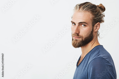Foto Close up of manly handsome guy with fashionable hairstyle and beard looking in camera, holding head in three quarters with serious expression