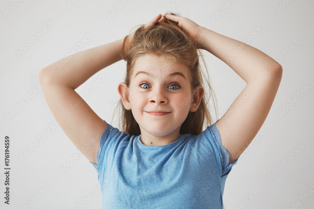 Funny cheerful blonde girl with blue eyes holding hair with hands, looking in camera with raised brows and silly face expression Funny cheerful blonde girl with blue eyes holding hair with hands, looking in camera with raised brows and silly face expression