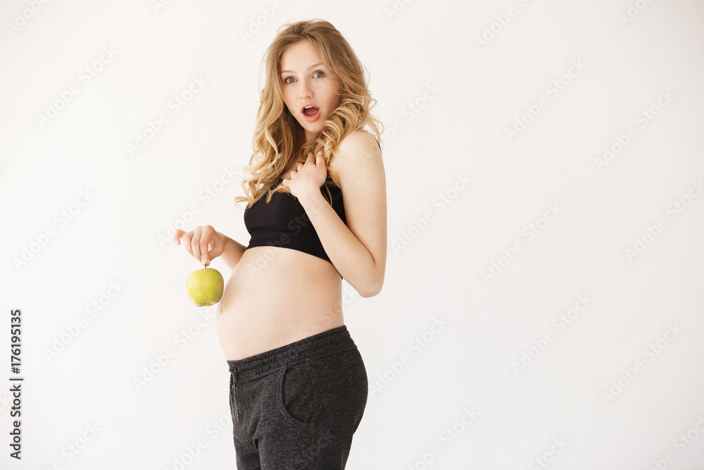 Close up portrait of good-ooking young european pregnant mother with blond hair looking in amera witth opened mouth and surprized face expression, holding apple near stomach. Woman funny posing for Close up portrait of good-ooking young european pregnant mother with blond hair looking in amera witth opened mouth and surprized face expression, holding apple near stomach. Woman funny posing for