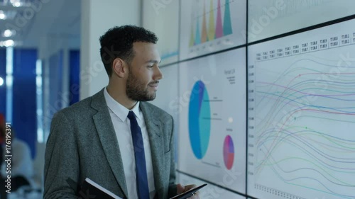  Portrait smiling businessman standing next to video wall with graphs & data