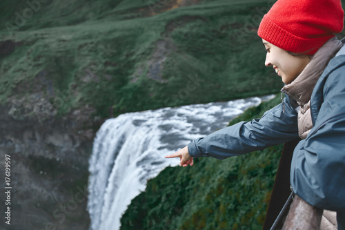 Girl in waterproof clothing stands on the cliff on background of Skogafoss waterfall in Iceland. View from above