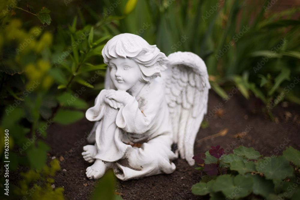 A small  white statue of a thinking angel in a garden between the plants.