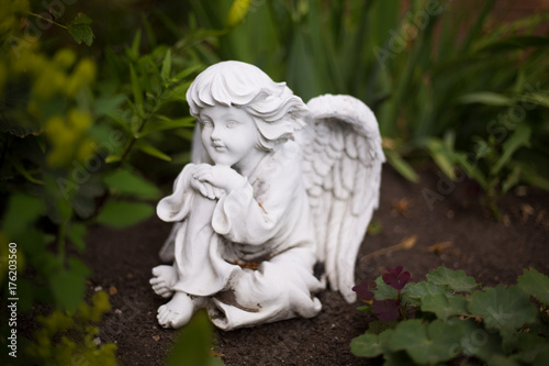 A small white statue of a thinking angel in a garden between the plants.
