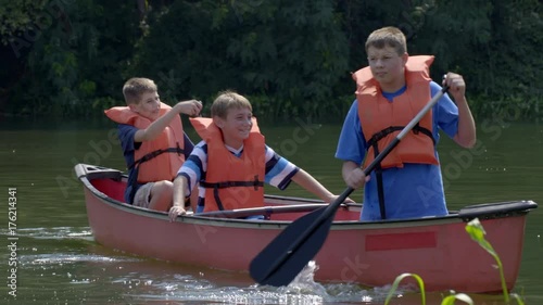 Three boys paddling in a canoe, turning towards camera on a large pond surrounded by trees. Close view in summer sunlight, 4K