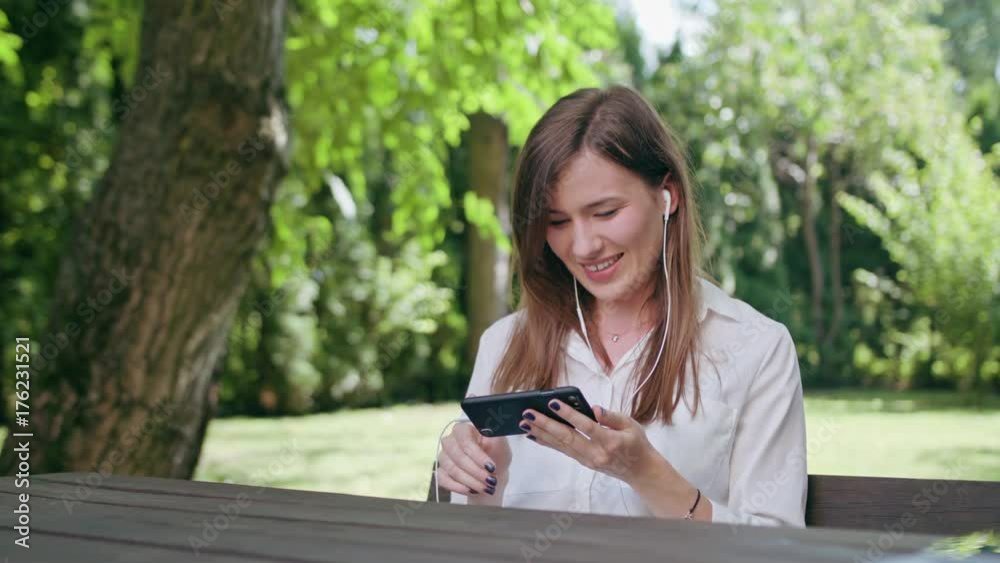 A young attractive lady sitting on the bench at the table in the park and listening to music using a phone. Medium shot. Soft Focus A young attractive lady sitting on the bench at the table in the park and listening to music using a phone. Medium shot. Soft Focus