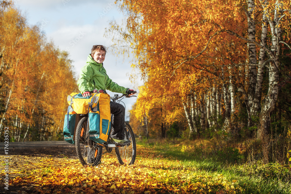 Obraz premium Young happy bicycle tourist with his loaded bike standing on an autumn road in a sunny day