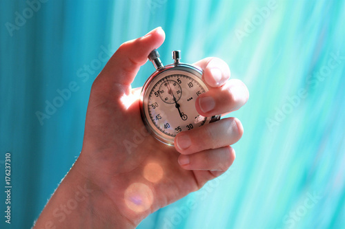 Hand holding a stopwatch and pressing the button to note the time. Sun rays flare and aquamarine background