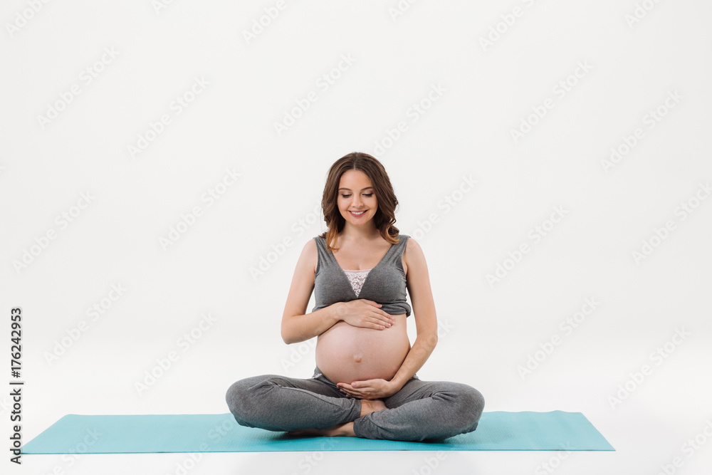 Smiling pregnant woman sitting on fitness mat and holding tummy Smiling pregnant woman sitting on fitness mat and holding tummy