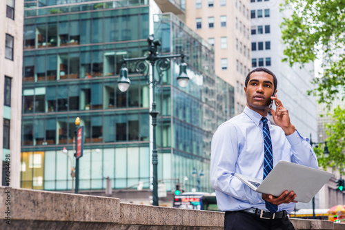 Obraz na płótnie Young African American Businessman working on laptop computer, talking on phone, standing by marble half wall on street in Manhattan, New York, wearing white shirt, blue pattern tie, looking away.. w salonie