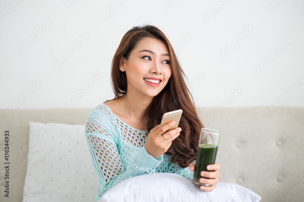 Smiling young asian woman drinking green fresh vegetable juice or smoothie from glass using smartphone at home Smiling young asian woman drinking green fresh vegetable juice or smoothie from glass using smartphone at home