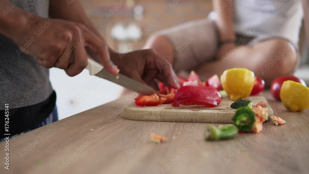 Closeup view of african guy's hands cutting vegetables preparing food while his caucasian girlfriend is sitting on the table in the kitchen in the morning. Happy multiethnic couple cooking breakfast Closeup view of african guy's hands cutting vegetables preparing food while his caucasian girlfriend is sitting on the table in the kitchen in the morning. Happy multiethnic couple cooking breakfast