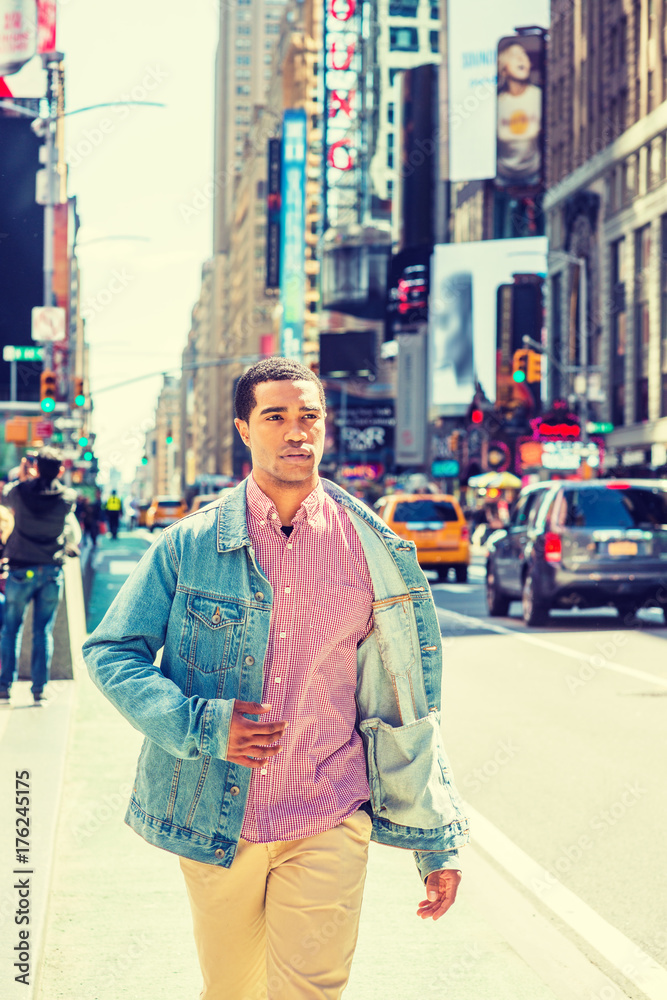 Young African American Man traveling in crowded Times Square, New York, wearing Denim jacket unbuttoned, red, white patterned shirt, yellowish brown pants, walking. High Buildings, cars on background.