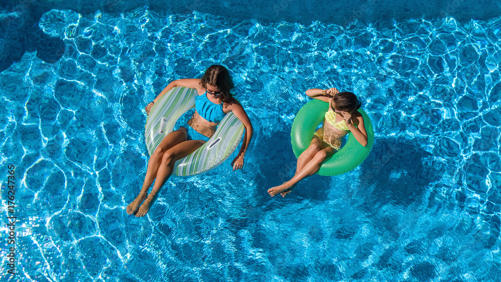Aerial top view of children in swimming pool from above, happy kids swim on inflatable ring donuts and have fun in water on family vacation

