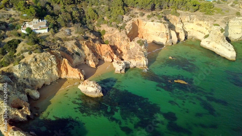 Shot taken with drone of green water washing sandy and rocky beach of coastline with house placed on hill above in bright sunlight, Portugal, Algarve.