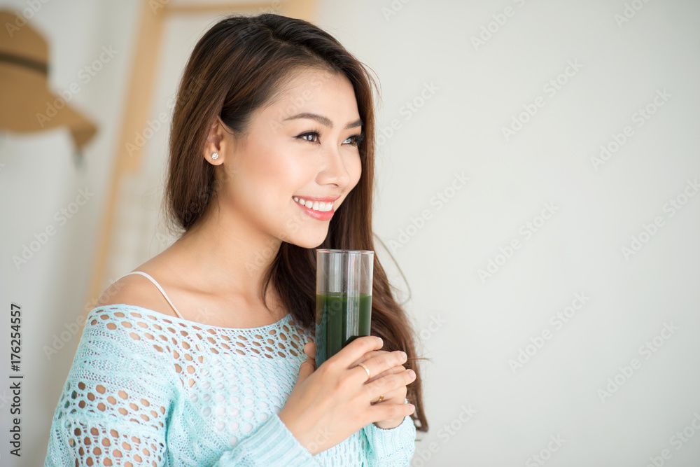 Smiling young asian woman drinking green fresh vegetable juice or smoothie from glass at home Smiling young asian woman drinking green fresh vegetable juice or smoothie from glass at home