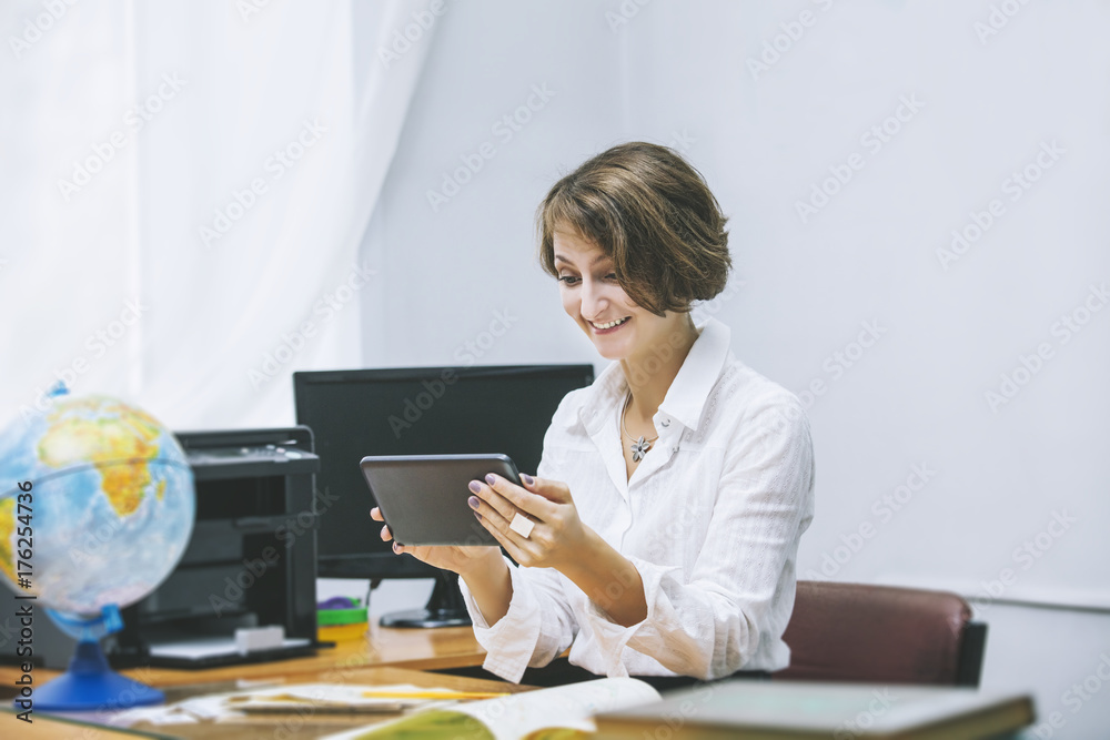 Teacher happy young woman in a classroom with electronic tablet in the workplace at the table Teacher happy young woman in a classroom with electronic tablet in the workplace at the table