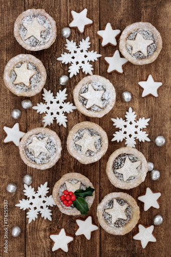 Mince pies and gingerbread biscuits with holly, snowflake bauble decorations and foil wrapped chocolates on oak wood background.