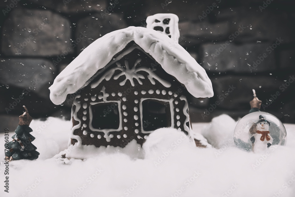 Naklejka premium Snowy Gingerbread house with snowflakes christmas tree and globe on stone wall background. Homemade christmas cookies - gingerbread on snow background. Closeup xmas gingerbread cottage in wintertime