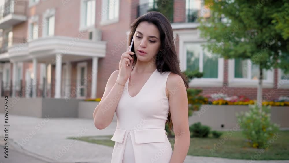 Young brunette in white talking smartphone and looking away while posing at street. Young brunette in white talking smartphone and looking away while posing at street.