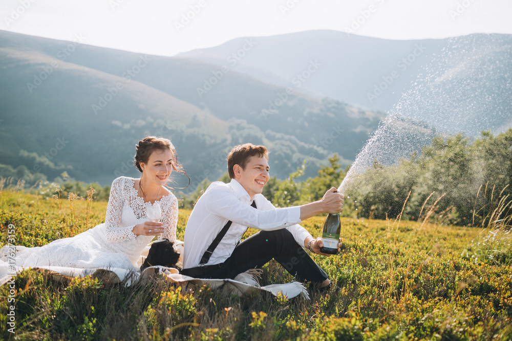 Bride and groom drink champagne sitting on a green hill