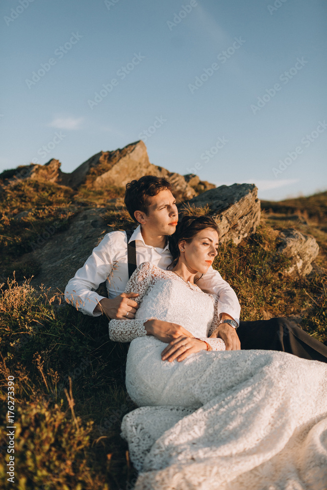 Cheerful newlyweds rest on the rocks in the rays of evening sun Cheerful newlyweds rest on the rocks in the rays of evening sun