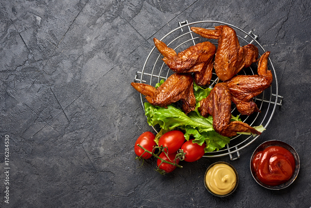 Smoked chicken wings on barbecue grill with tomatoes, mustard and ketchup. Black concrete background. Top view. Space for text.