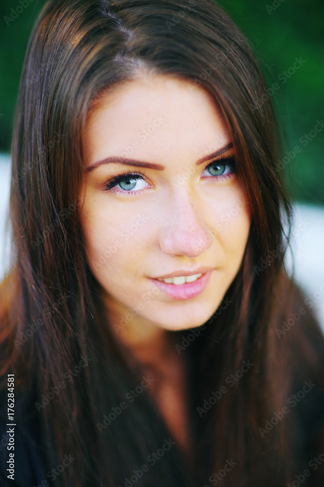 Naklejka premium Portrait of a beautiful young blue-eyed girl with a cute sincere smile in the rays of the setting sun on a blurred background of green leaves in summer outdoors