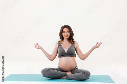 Smiling pregnant woman sitting on fitness mat and meditation