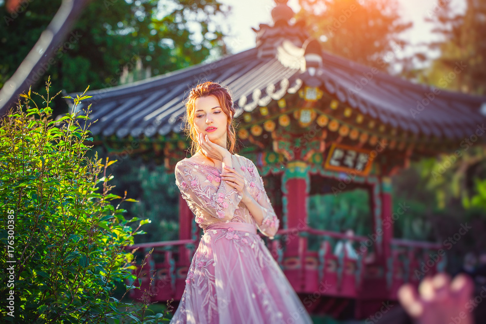 Fototapeta premium Beautiful girl with red hair in a pink dress. The background of Japanese architecture in the garden. Spring garden.