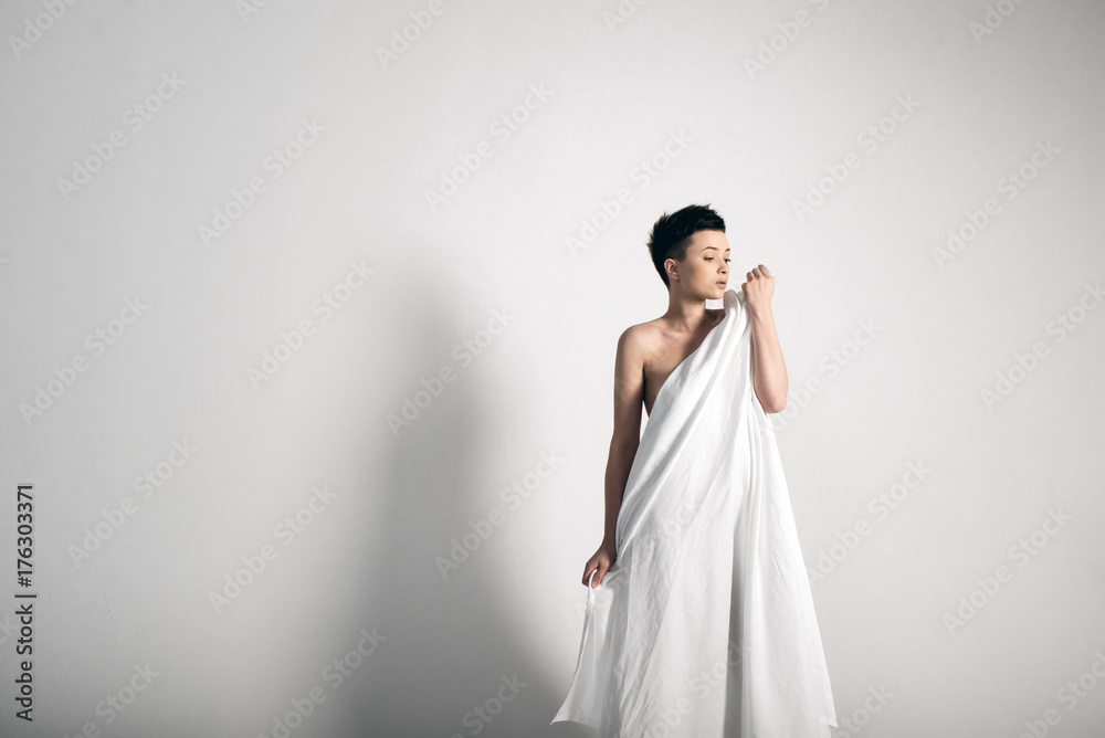 Fototapeta premium Sensual naked young adult caucasian woman, wrapped in a satin, silk sheet against grey background. High contrast
