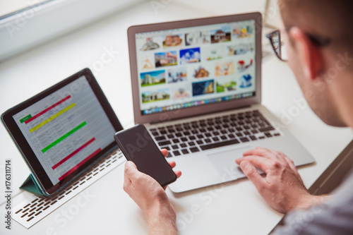 Close-up young man using the laptop and keeping the phone in hand indoors