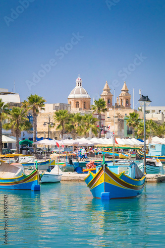 Traditional colourful Maltese Luzzu fishing boats in the turquoise blue water of Marsaxlokk harbour, with the beautiful Parish Church of Our Lady of Pompei, Marsaxlokk, Malta, June 2017 photo