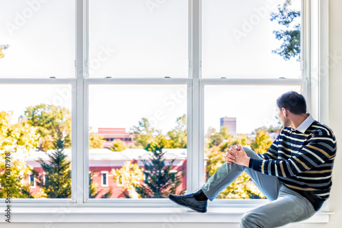Tableau sur toile Young man sitting on windowsill by large window overlooking red brick buildings