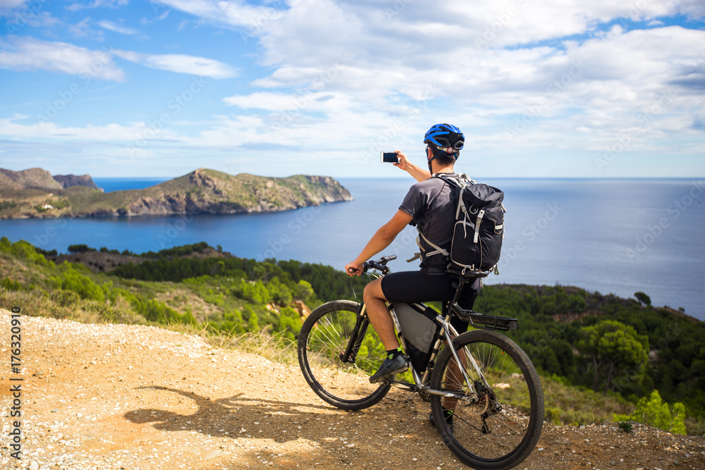 a young guy on a mountain bike trails in Spain and takes a photo on a white phone in the background of the Mediterranean sea of the rocky coast of the Costa Brava. 