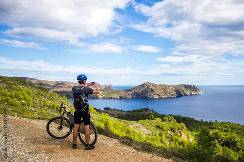 a young guy on a mountain bike trails in Spain and takes a photo on a white phone in the background of the Mediterranean sea of the rocky coast of the Costa Brava Fototapeta