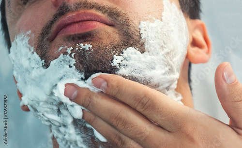 A young man is shaving in the morning, smiling. On the face there is a lot of foam for shaving. Toning