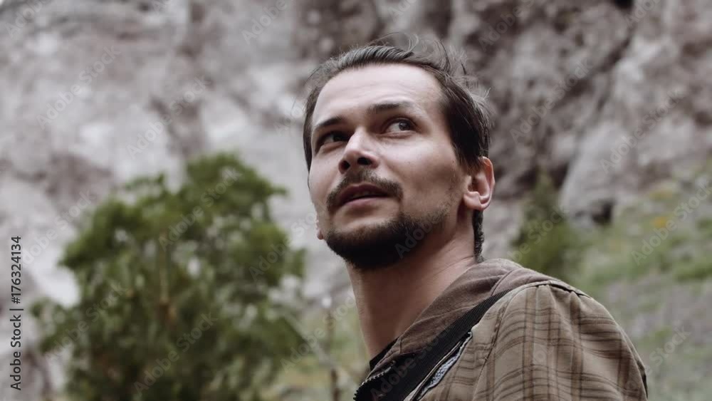 Young bearded caucasian man in grey jacket at mountain hiking trail standing near waterfall at windy day Young bearded caucasian man in grey jacket at mountain hiking trail standing near waterfall at windy day