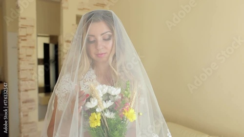 Beautiful young bride in a wedding veil with a covered head is standing with a bouquet of flowers near the window. photo