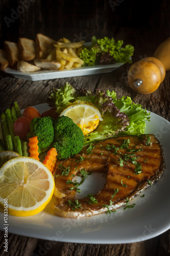 Salmon steak with sauce on a white plate with lemon on plate and crispy French fries. Many vegetables are placed around the dish on the wooden floor.